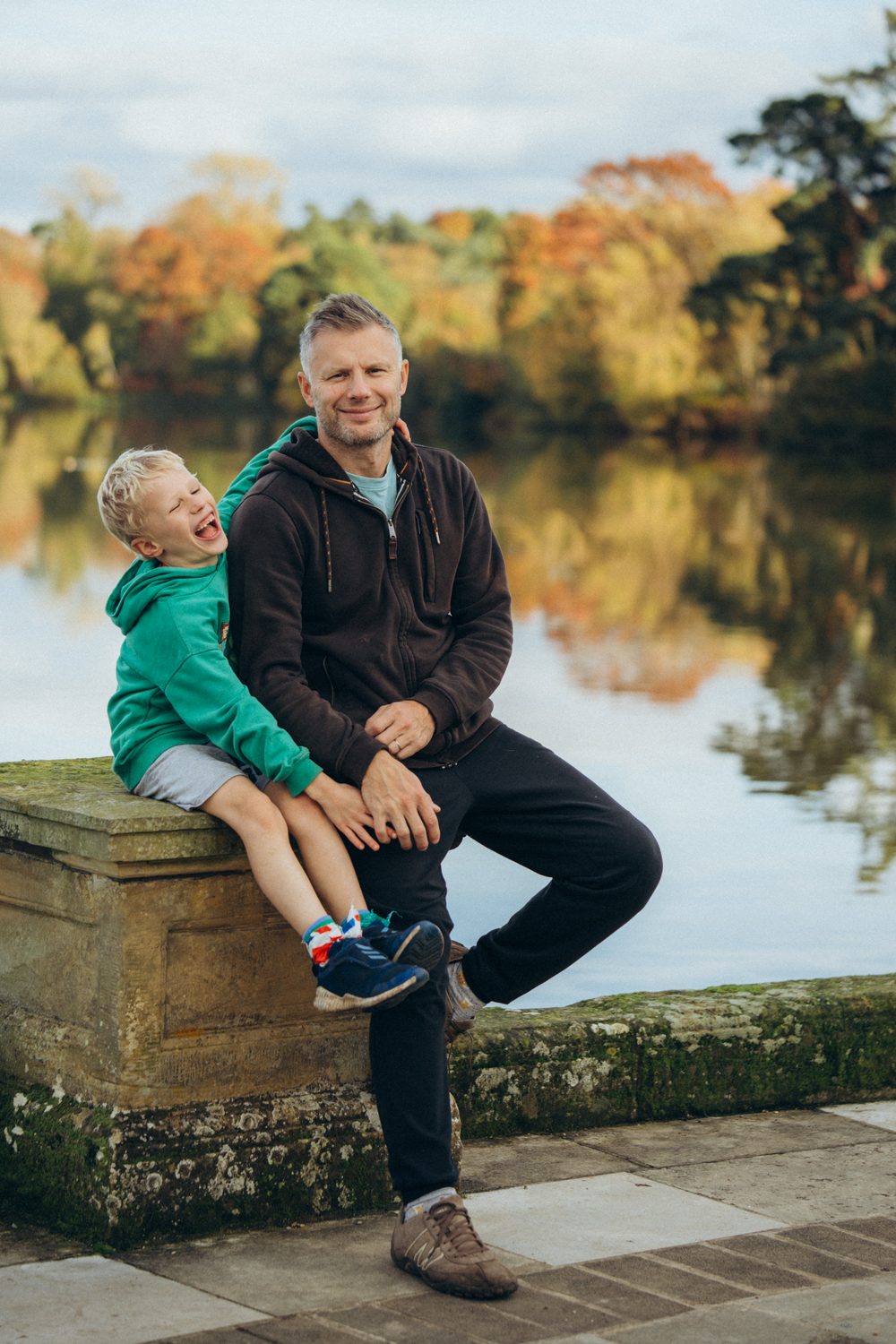 Father and son laughing together by a lake on an autumn family shoot