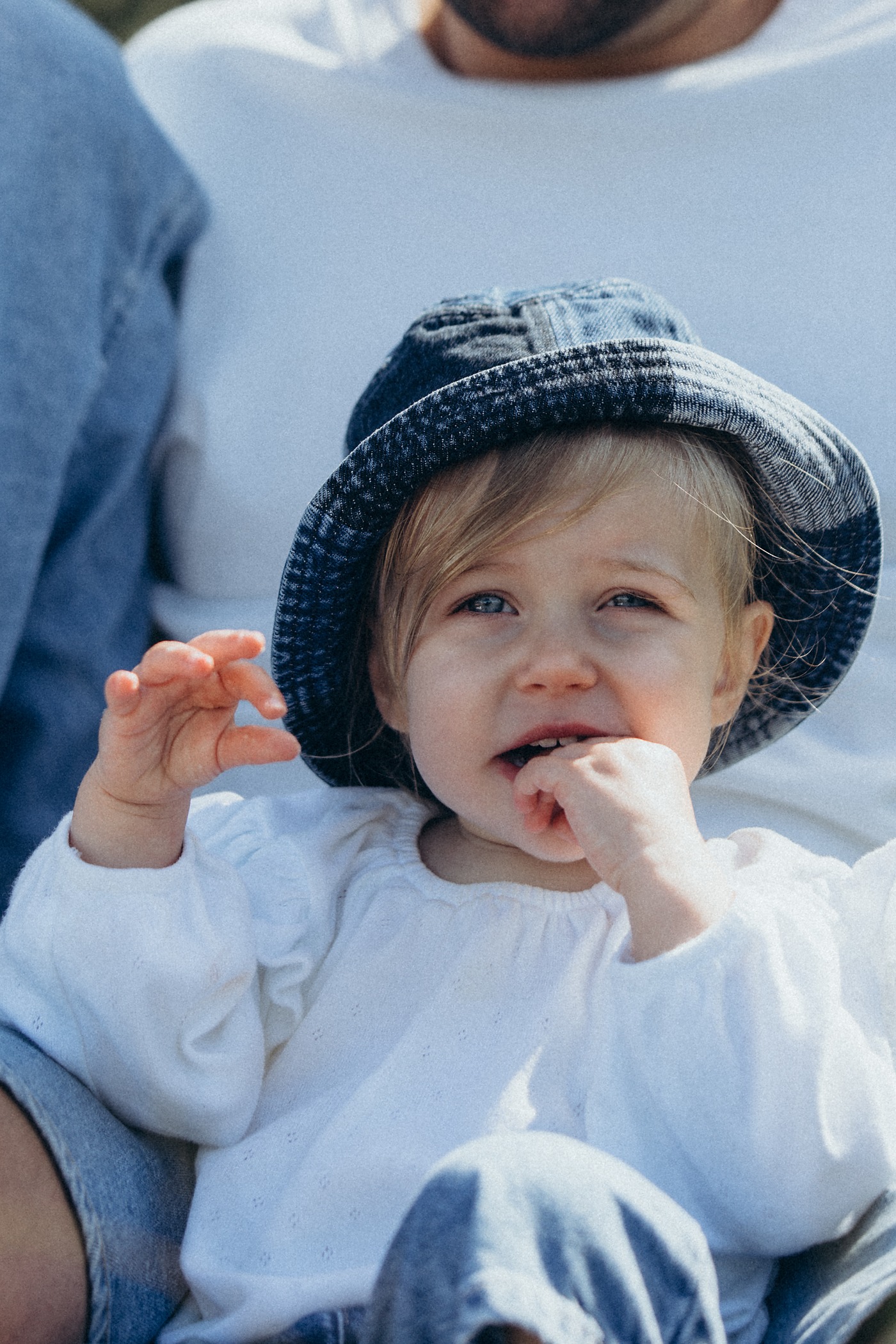 Toddler in a denim bucket hat smiling with her fingers in her mouth