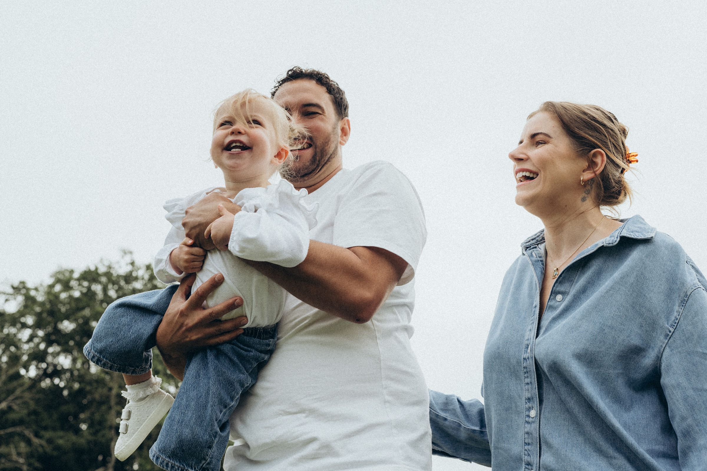 Father lifting his laughing toddler into the air while her mother watches, in a summer meadow