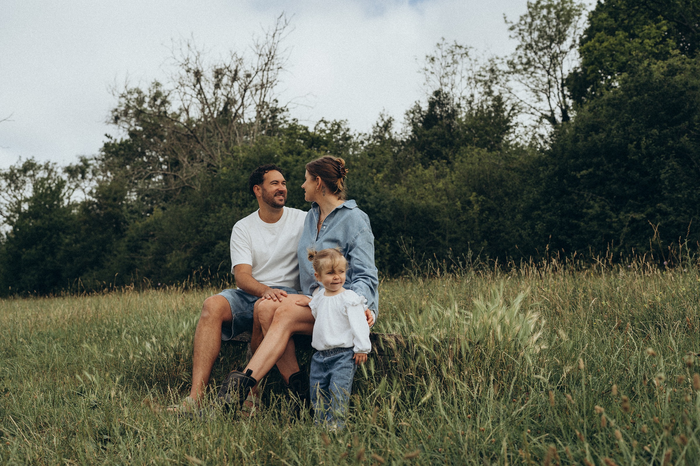 Family of three sitting together in a summer meadow with trees behind them