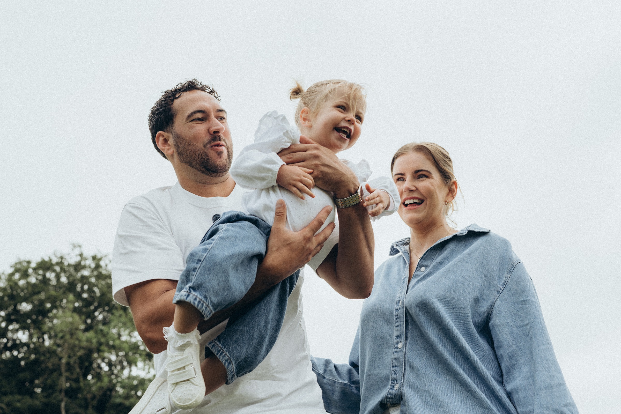 Parents in denim and white lifting their toddler between them in a meadow