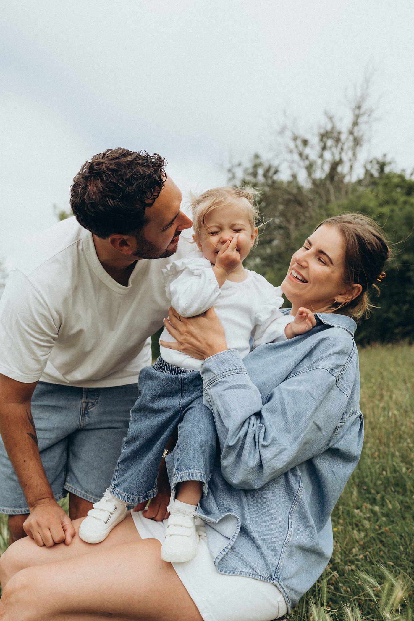 Dad kissing his toddler while mum laughs, kneeling in the grass
