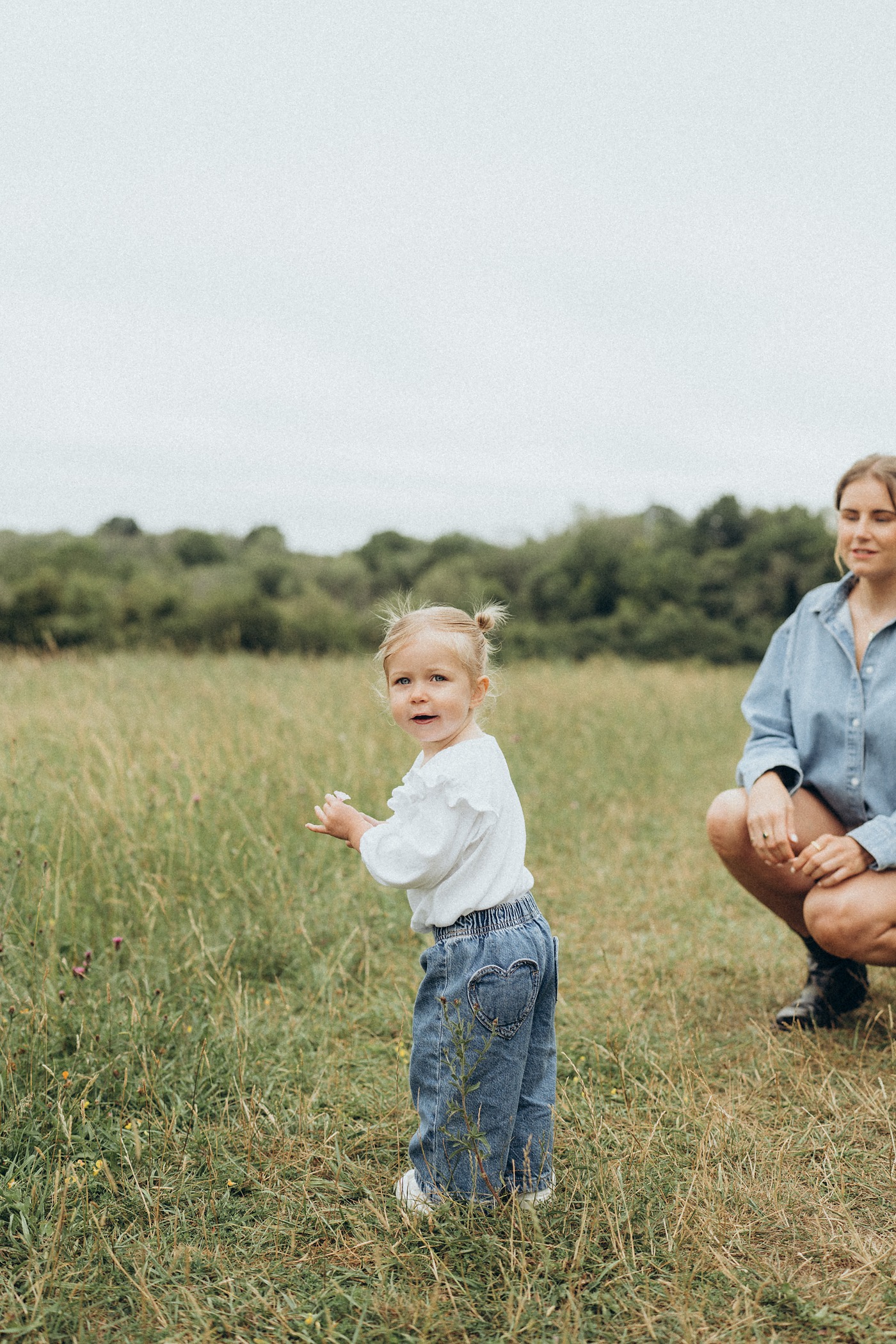Toddler walking through a meadow as her mum crouches nearby