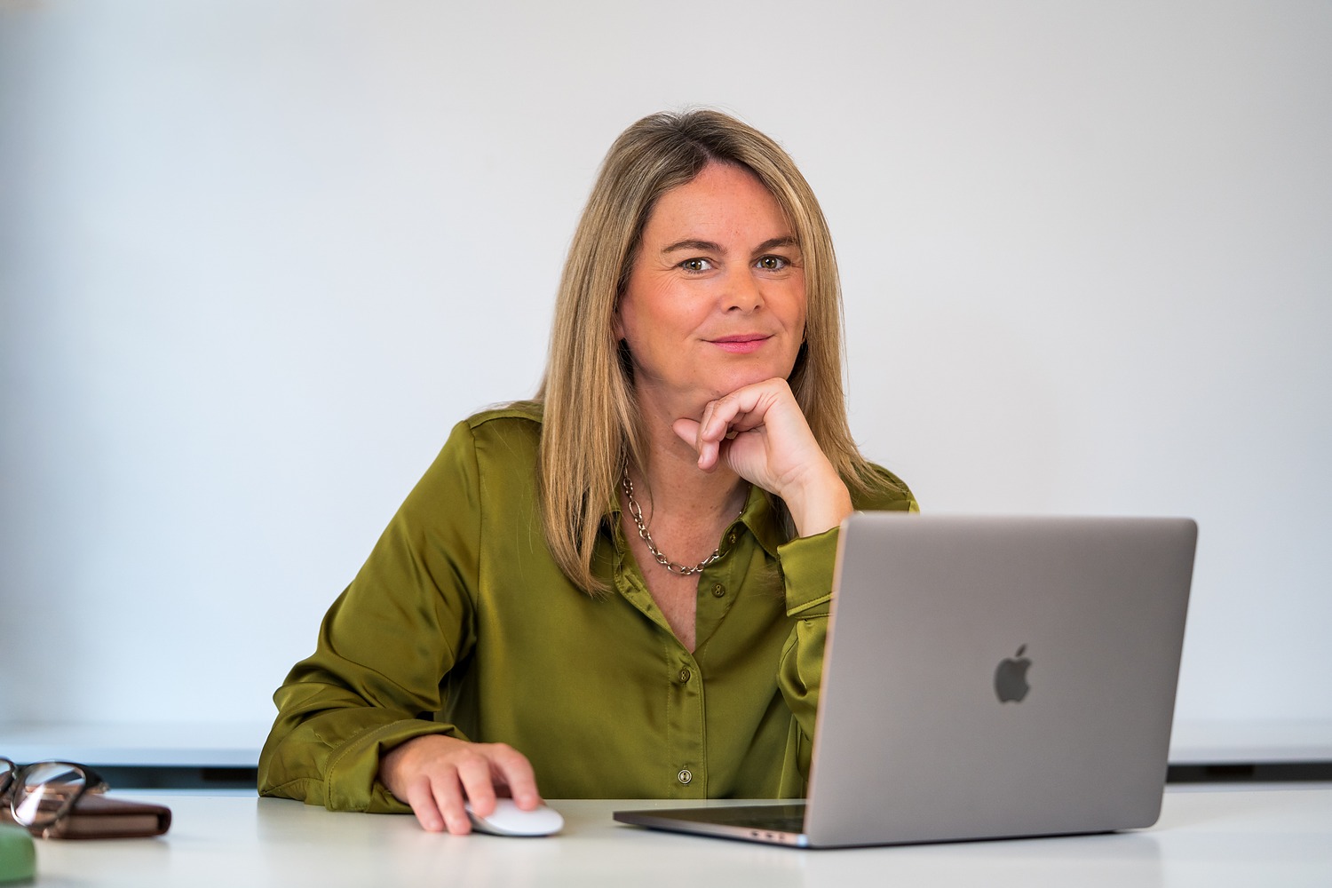 Relaxed business portrait of a woman in an olive silk shirt at her desk, chin resting on her hand beside her laptop