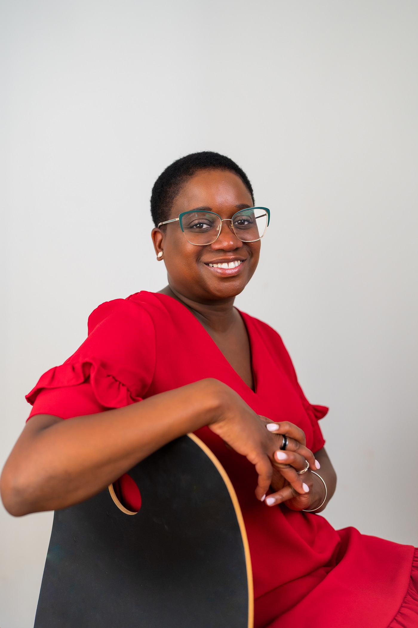 Woman in a red dress and teal glasses smiling over the back of a black chair in a relaxed headshot