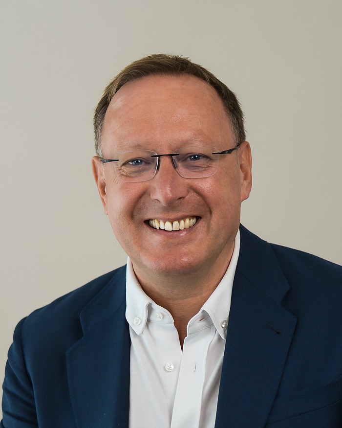Headshot of a smiling man in glasses and a navy blazer against a pale grey backdrop
