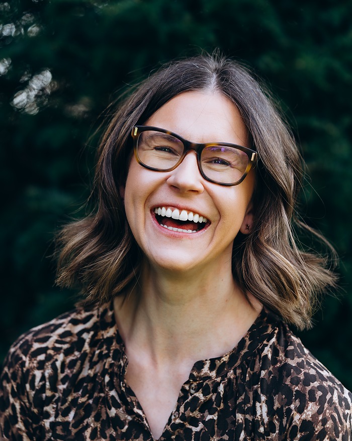 Headshot of a woman in glasses laughing, against dark green leaves