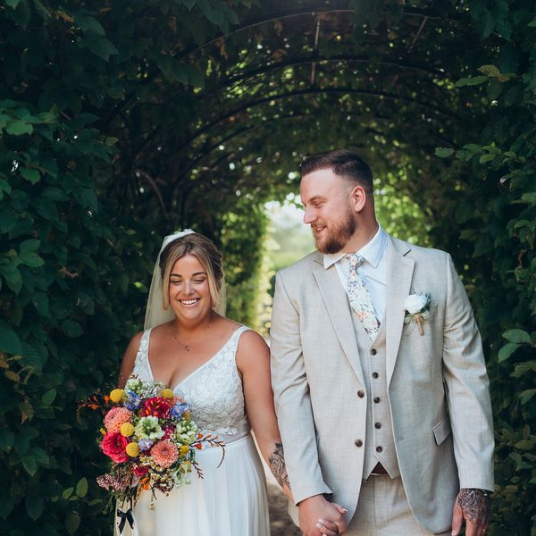 Bride and groom side by side under an arch of trees, her bouquet in hand
