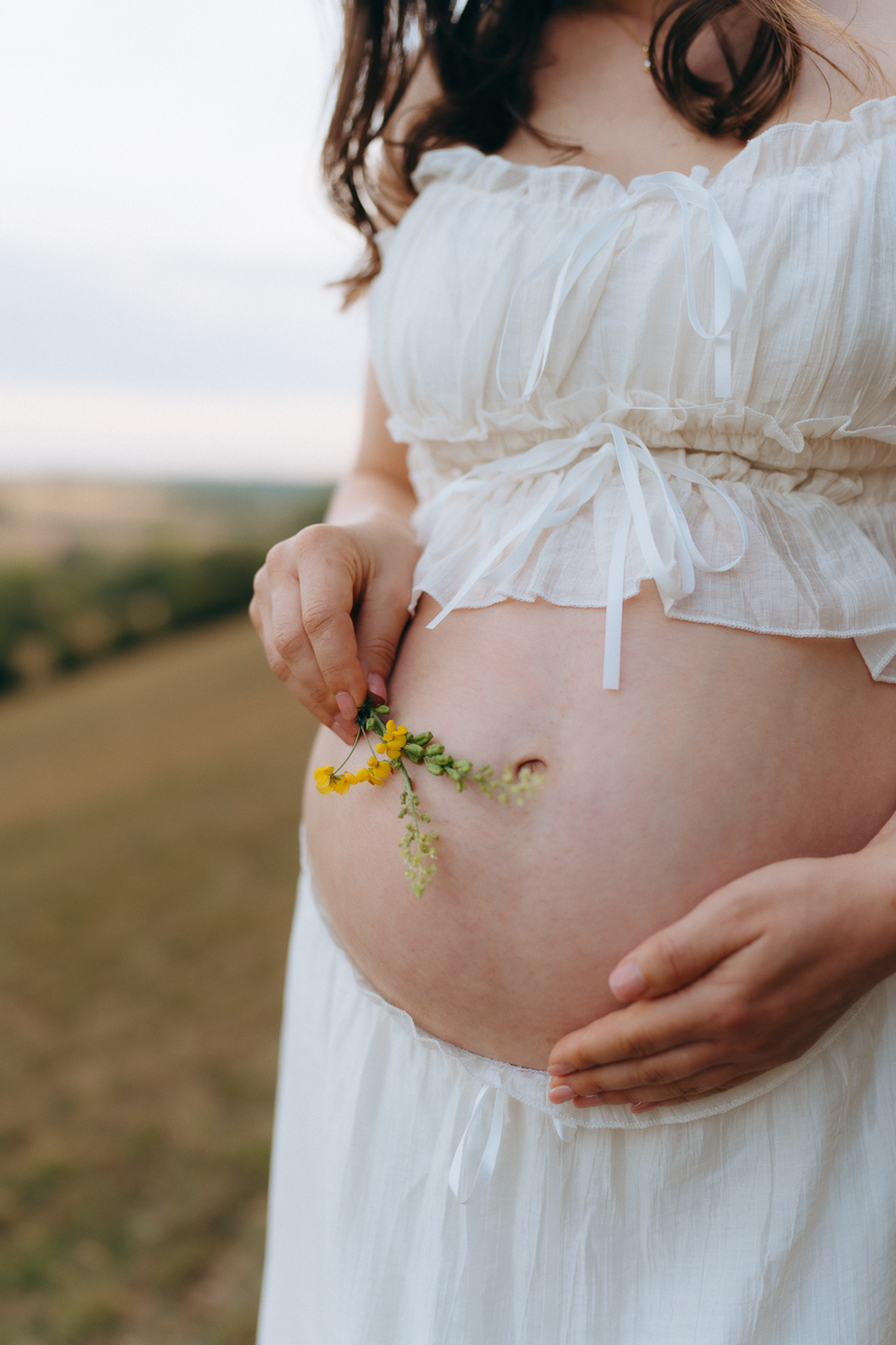 Mother-to-be holding yellow wildflowers by her bump on a hillside in the Surrey countryside