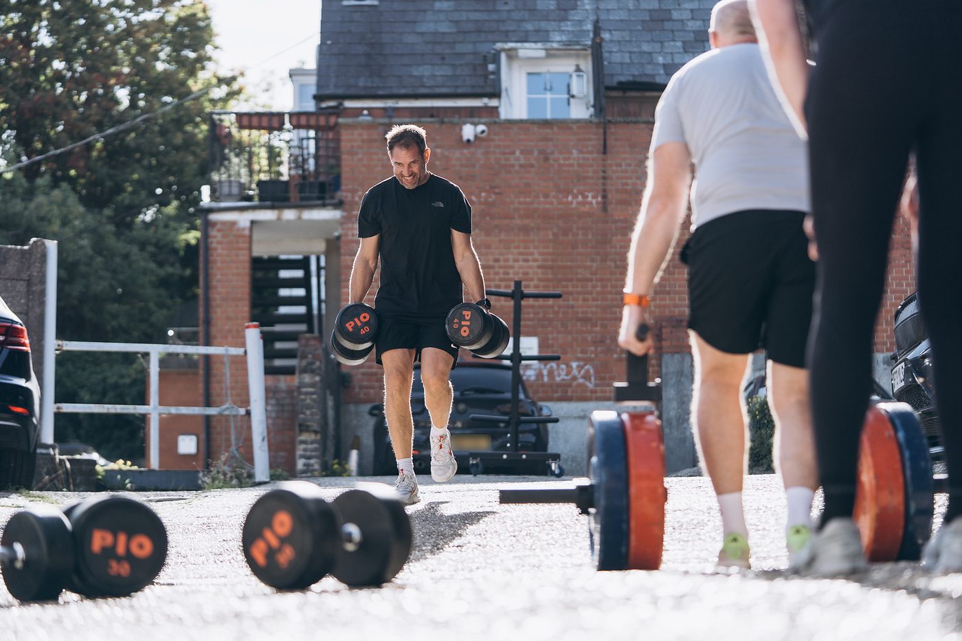 Member carrying two heavy dumbbells across the yard outside the gym