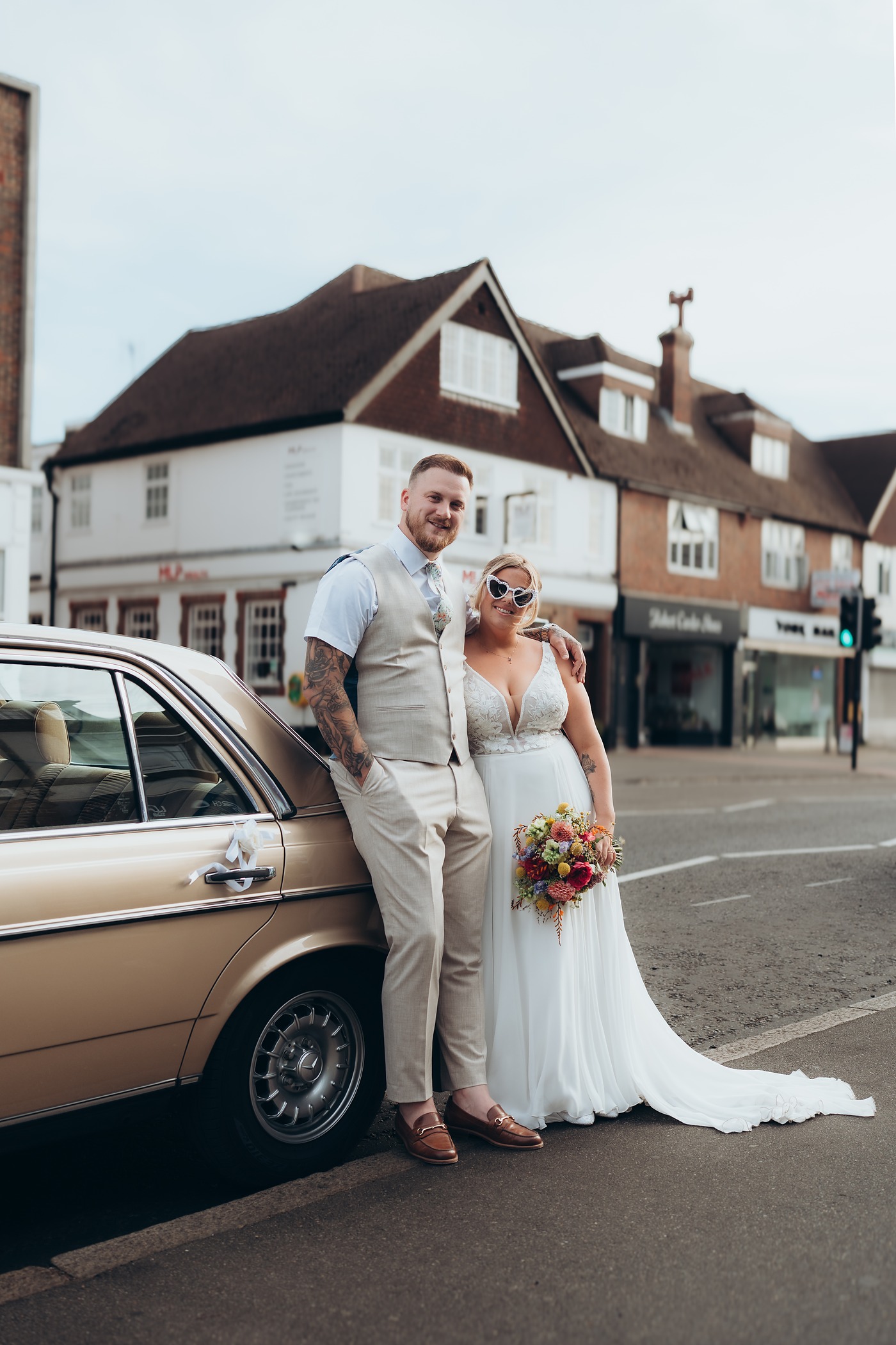 Bride in heart-shaped sunglasses and her groom leaning on a vintage car in the high street