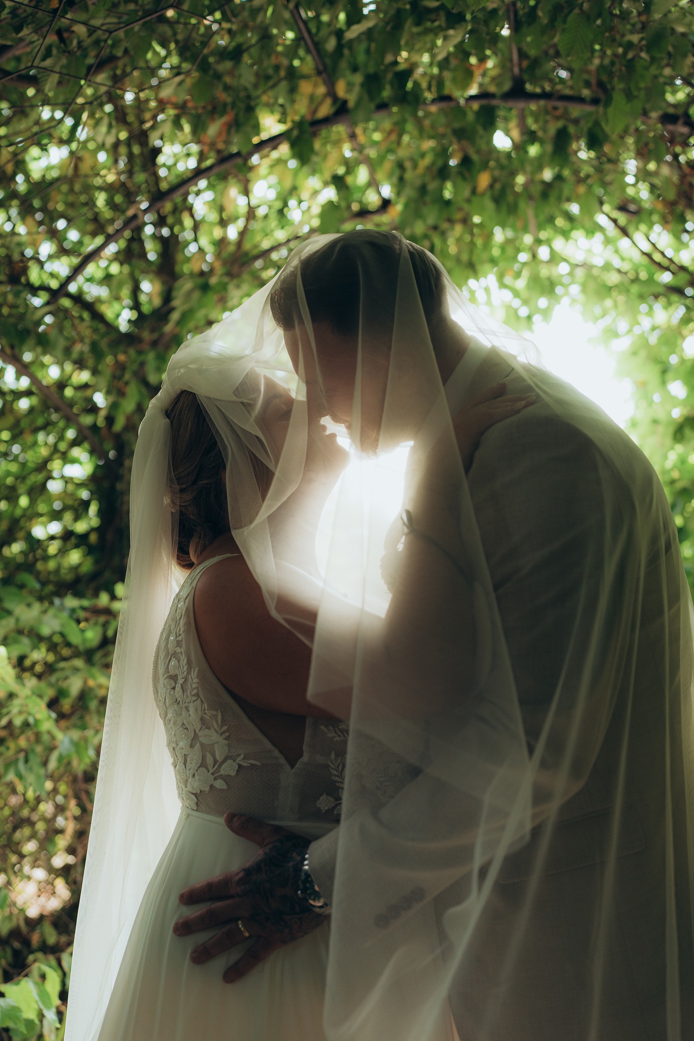 Bride and groom kissing beneath her veil, lit from behind by the sun through the trees