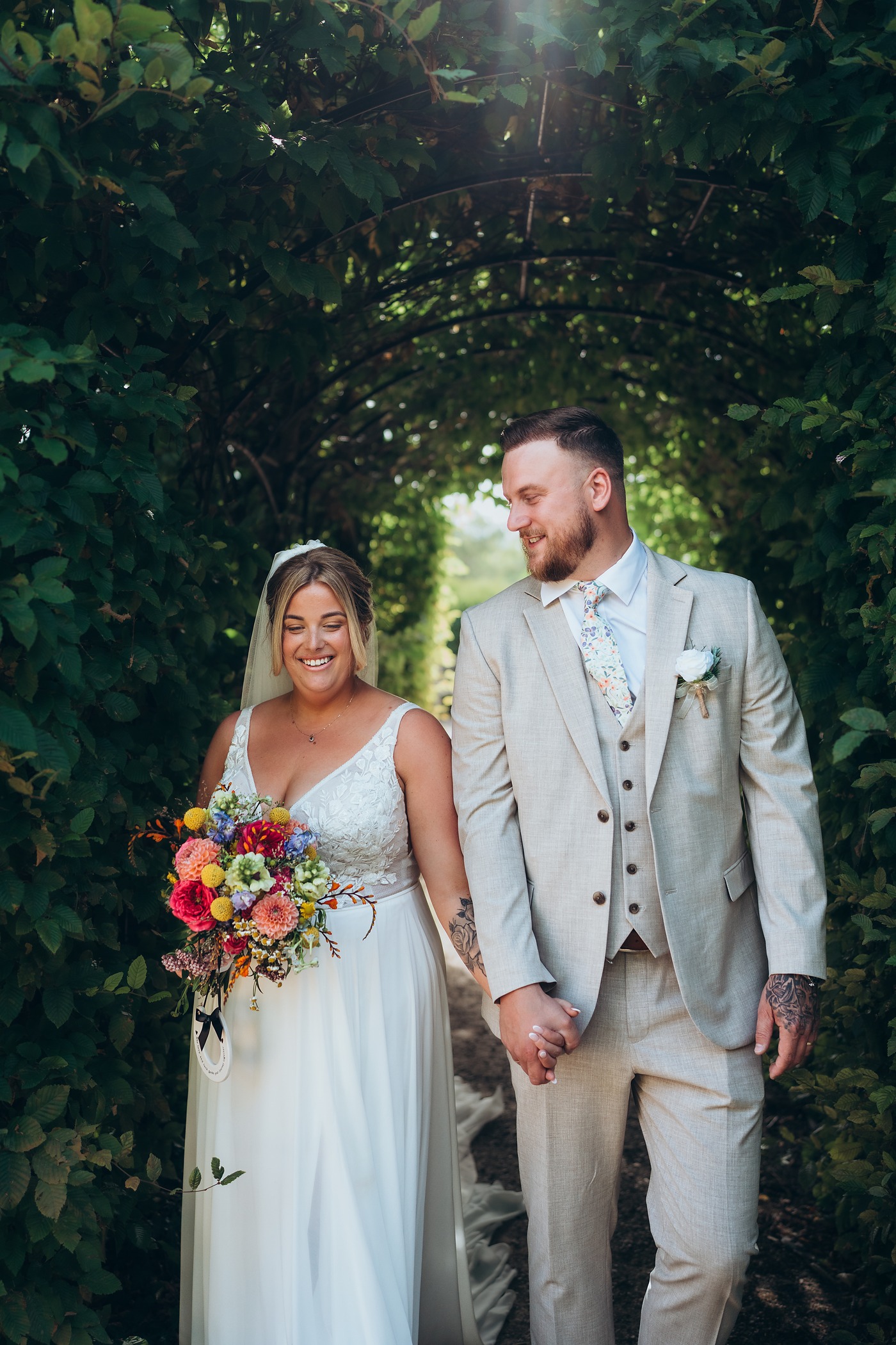 Bride and groom walking hand in hand through a leafy archway, her bouquet full of colour