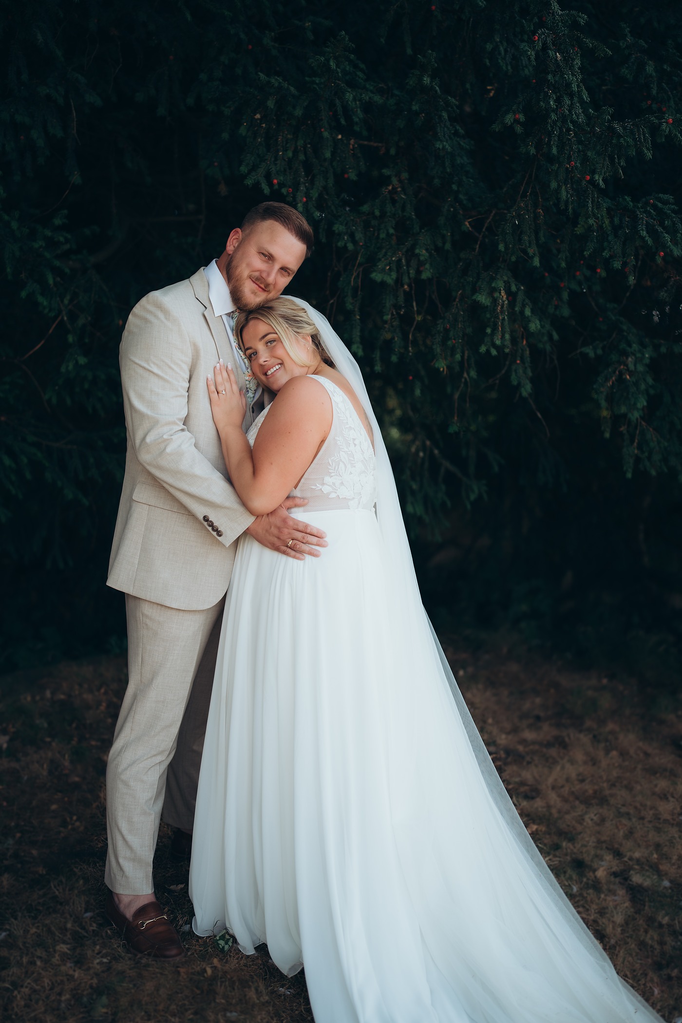 Bride resting her head on her husband's chest in front of a dark yew tree